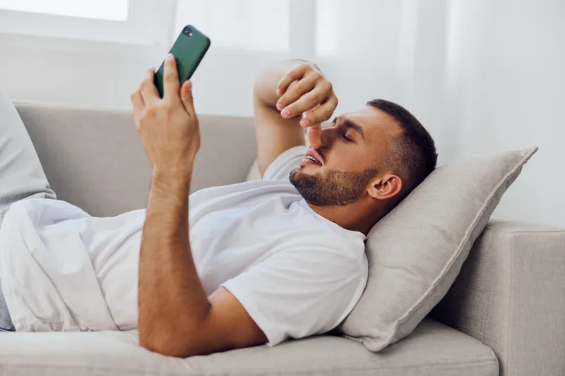 Relaxed man lying on a sofa while using a smartphone, showcasing emotions of contemplation and leisure. A soft, modern living space enhances the vibe.