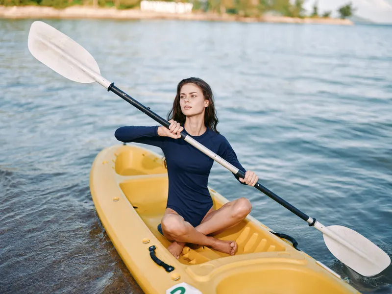 A young woman sits cross-legged in a yellow kayak, paddling on calm blue water with a paddle raised high, showcasing balance, focus, and adventurous summer activity.