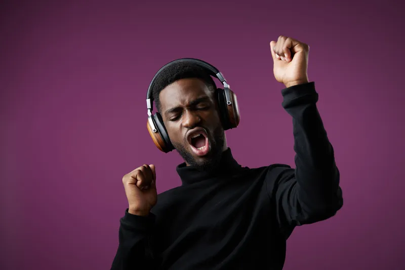 Young Black man listening to music with headphones, dancing joyfully against a purple background, expressing happiness through dynamic gestures