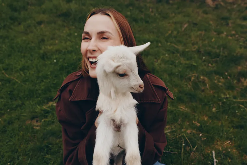A young woman with long brown hair joyfully holds a white baby goat outdoors on green grass during a casual lifestyle moment in nature.