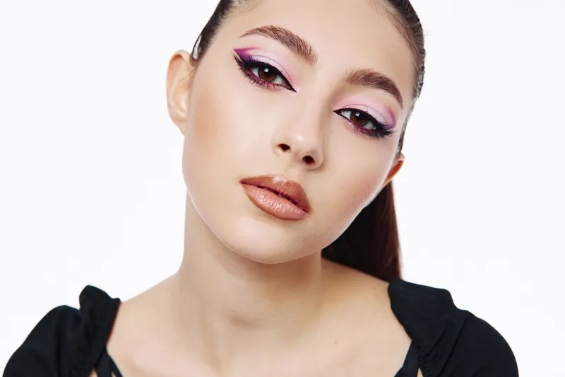 Young woman with creative pink and black eye makeup looks confidently at the camera. She wears a black top on a clean white background. Concept of beauty, fashion, and artistic makeup.