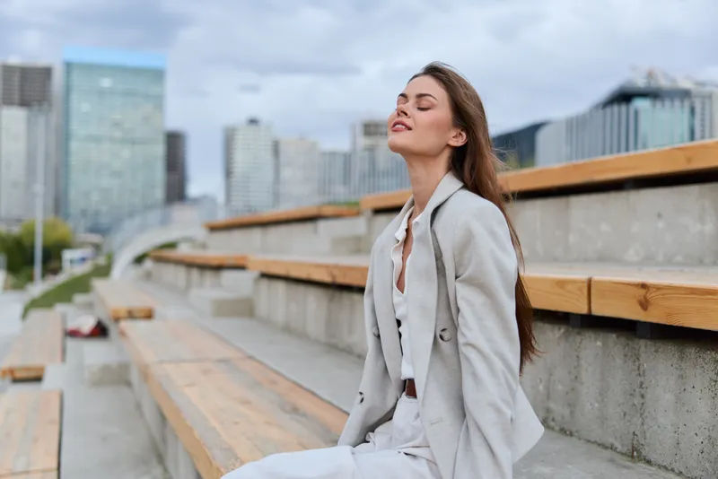 Confident businesswoman enjoying a moment of reflection outdoors in a modern cityscape, embodying professionalism and elegance in neutral attire.