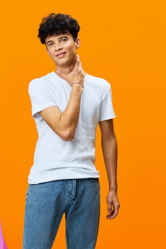 Young man standing in casual white t-shirt and jeans against vibrant orange background, with relaxed pose and thoughtful expression, natural lighting bright atmosphere.