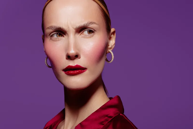Close-up of a serious woman with red lipstick and hoop earrings wearing a red satin blouse against a purple background. Concept of fashion, beauty, and confident expression.