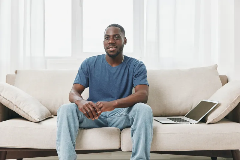 Smiling young African man sitting comfortably on a sofa at home, showcasing casual fashion and a relaxed atmosphere, ideal for lifestyle and wellness concepts