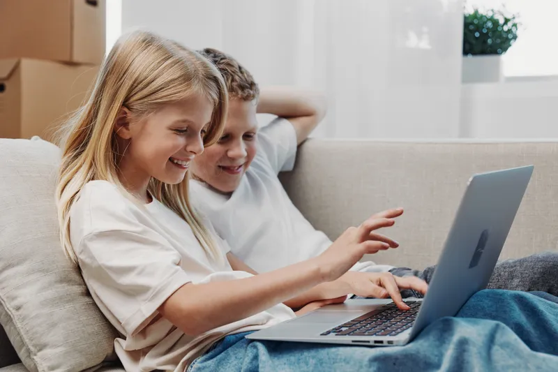 Happy children sitting on couch using laptop together, smiling and pointing at screen. Bright modern room with natural light. Concept for learning, technology and childhood development.