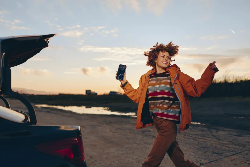 Woman lifestyle outdoors at sunset beside a car, holding a phone and smiling, curly hair, casual jacket and jeans, golden light and road scene, travel and connection concept
