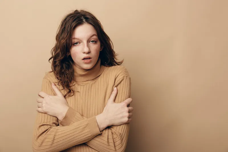 Tired young woman in brown sweater hugs herself with closed eyes, standing against beige background. Concept of fatigue, cold, discomfort, and emotional exhaustion in casual clothing.