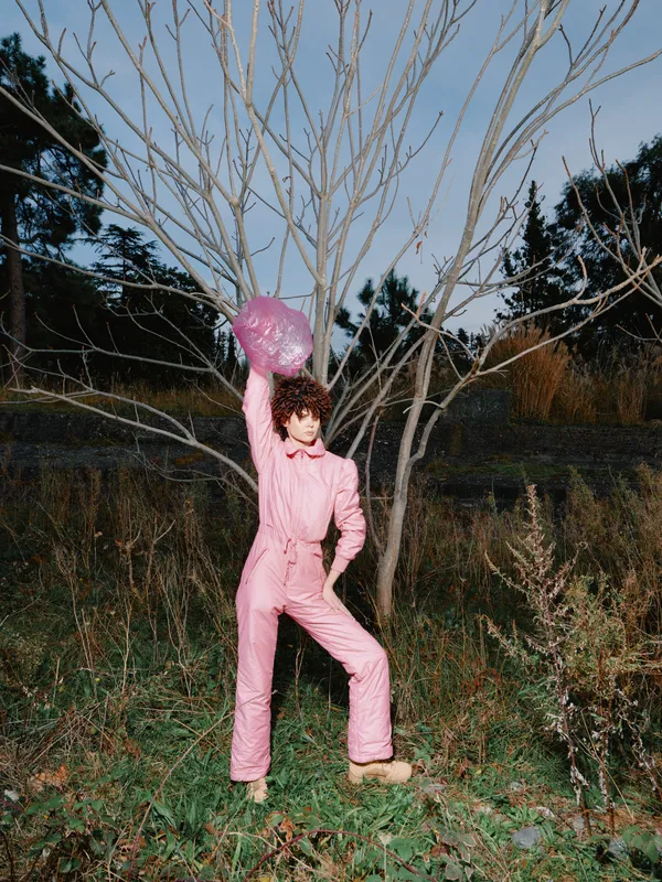 Person wearing a pink jumpsuit and matching helmet stands outdoors beside a leafless tree, striking a bold pose in a dusk-lit natural setting with tall grasses, wild bushes, and a muted sky above