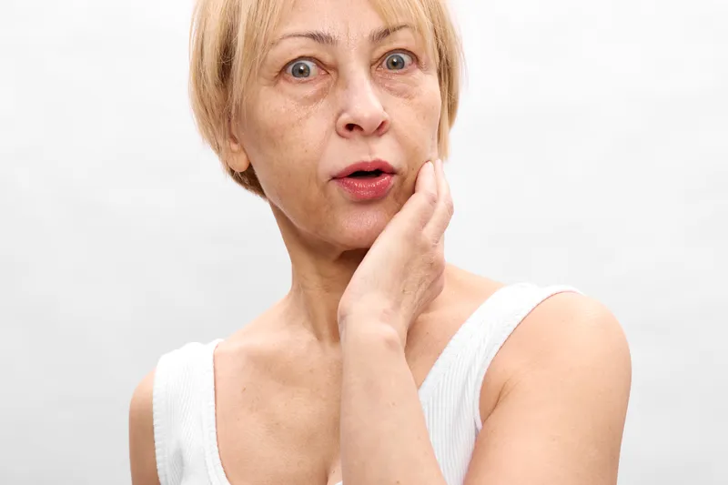 Elderly woman shows stress and worry, hand on cheek, surprised face, in a bright white studio portrait, emotional moment suggesting anxiety and burnout, today