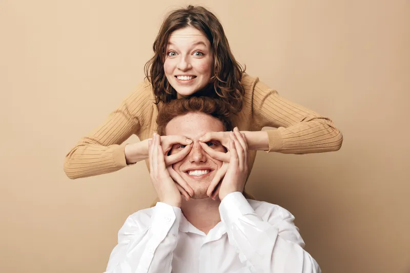 Happy young couple having fun together, woman playfully covering man's eyes with her hands. Both smiling and enjoying light moment in casual clothes on beige background.