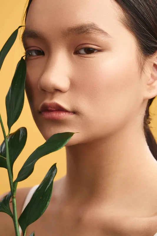 An asian woman is photographed in a soft studio setting, holding a leafy plant near her face. Calm expression, clear skin, gentle lighting convey serenity and quiet confidence.