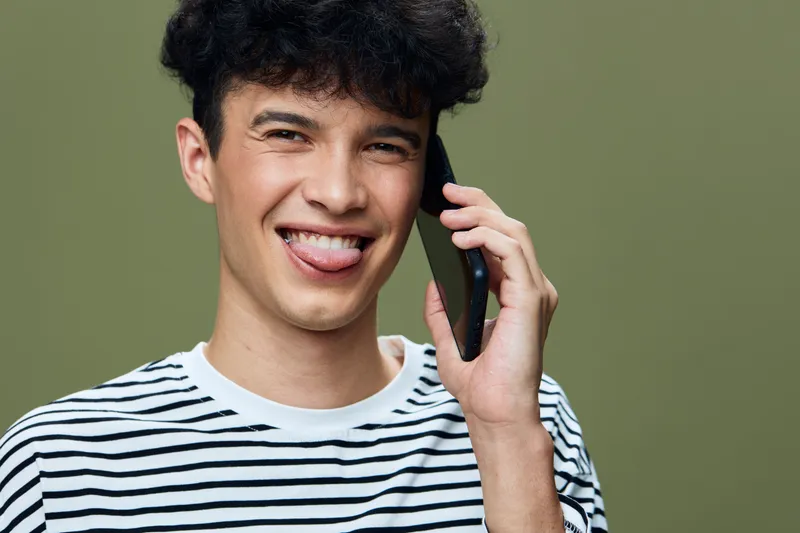 smiling man speaking on smartphone showing playful expression with tongue out, wearing striped shirt against green background, casual young male portrait