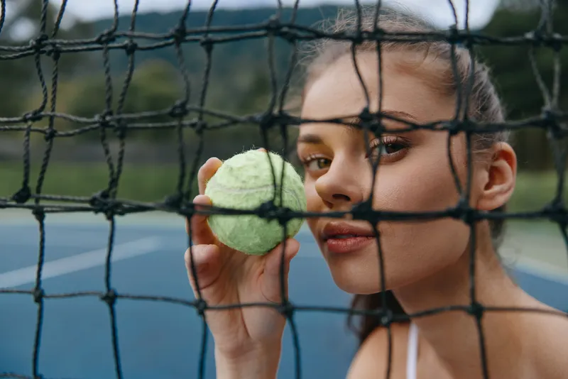 Young woman holding a tennis ball, expressing determination on a tennis court, surrounded by nature, highlighting sports enthusiasm and active lifestyle
