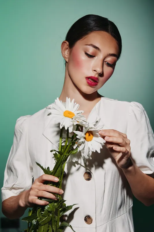 Young elegant woman in white dress with bright makeup delicately holds a bouquet of white daisies, posing thoughtfully against a soft green background. Beauty fashion concept.