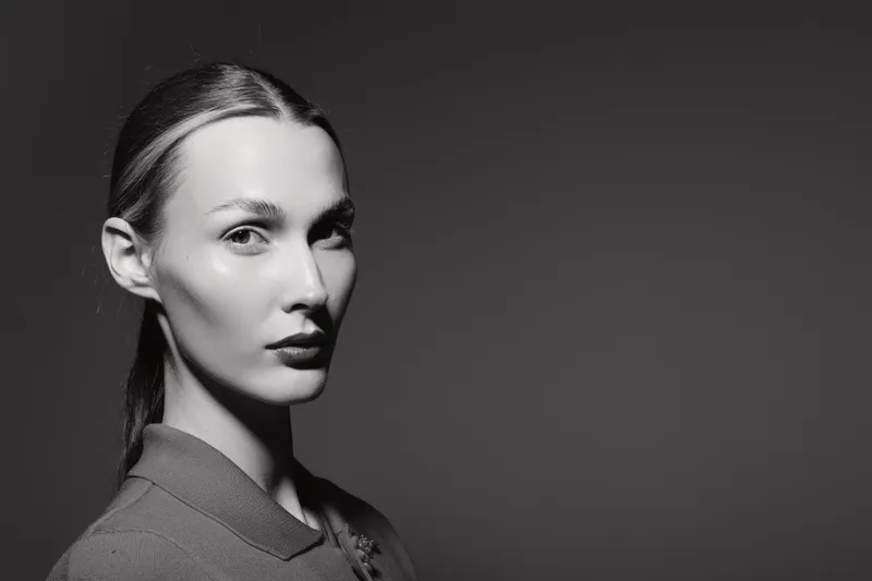 Portrait of a young white woman with pale skin and slicked back hair in studio. The monochrome image shows a serious facial expression with soft lighting on gray background.
