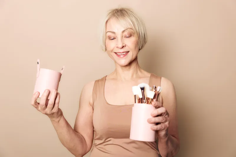 Mature woman with short blonde hair holds pink containers filled with makeup brushes against beige background. Concept of beauty routine, self-care, and cosmetic tools for skincare.