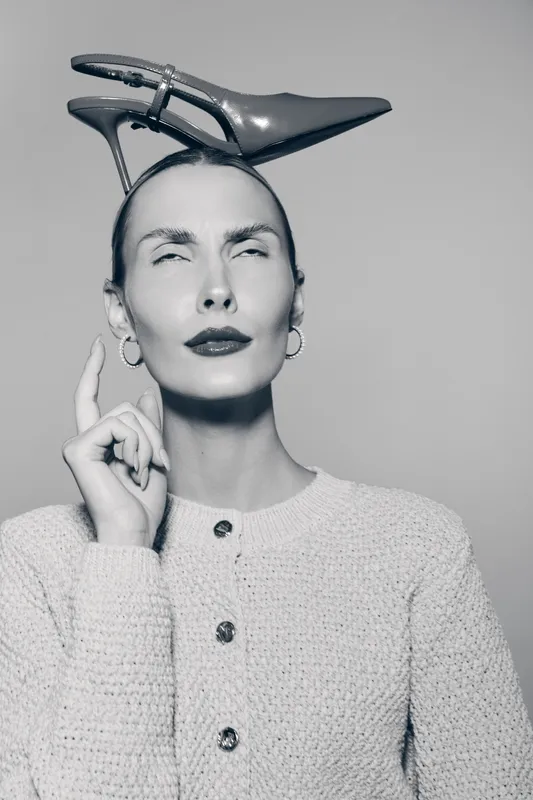 Elegant white woman with pale skin balances shiny high heel shoe on head, wearing knitted sweater and earrings in studio.