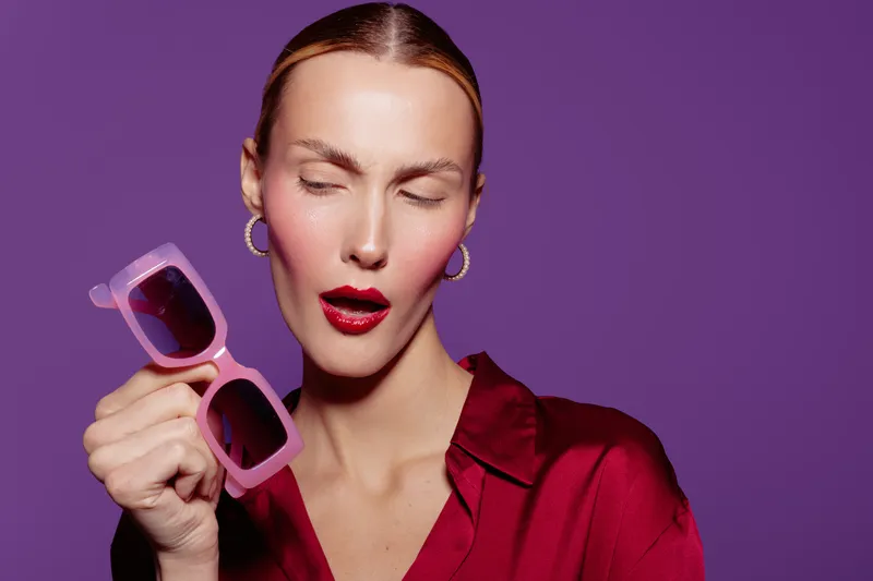 Young woman with closed eyes holds pink sunglasses in hand against purple background. She wears red lipstick and red blouse, expressing playful and stylish mood in studio lighting.