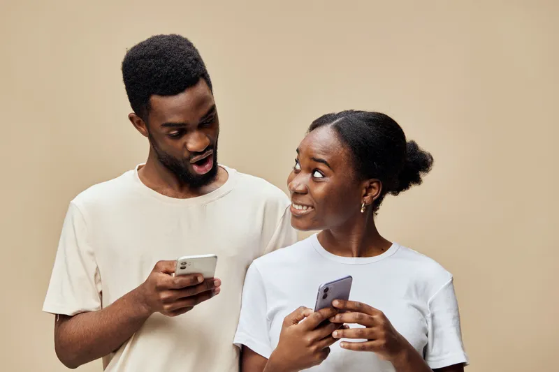 Young African couple enjoying conversation while using smartphones with warm background, showcasing happiness and connection through technology