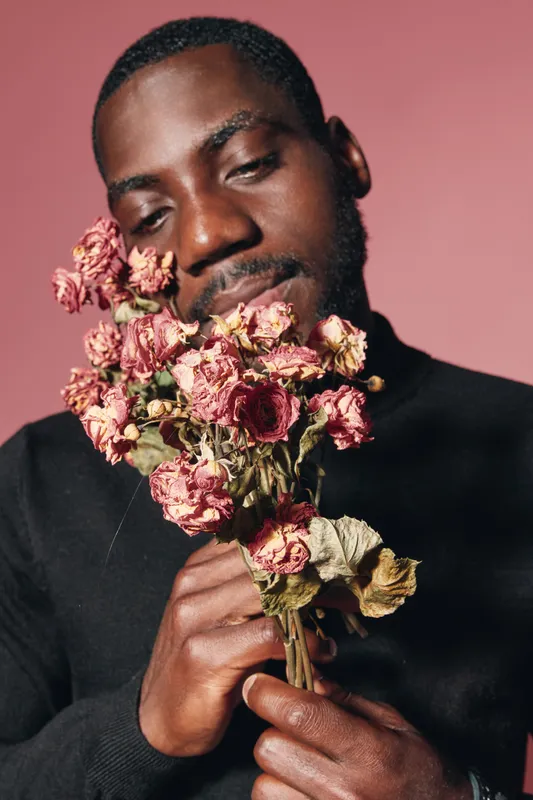 African man holding dried pink roses close to face with closed eyes in creative conceptual visual style. Portrait with emotional expression and dark skin on pink background.