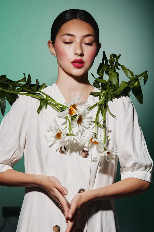 Young woman with bright red lips and flower decorations posed in a white dress against a vivid green background. Concept for beauty, fashion, and creative makeup.