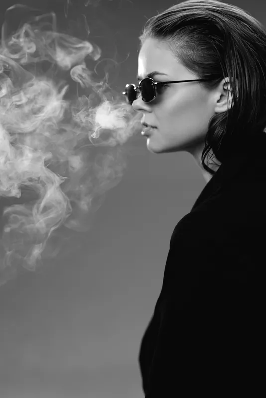Black and white portrait of a young woman wearing sunglasses exhaling smoke in a dark background.