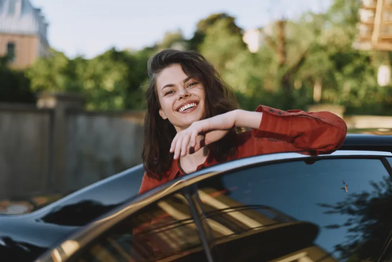 Woman lifestyle moment leaning on a car smiling at the camera in a sunny outdoor setting, showing casual fashion and urban leisure with confidence