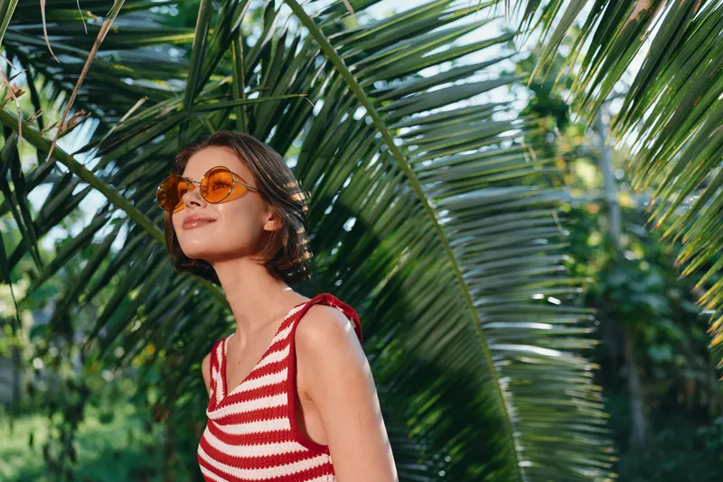 A cheerful woman wearing a red and white striped sleeveless top stands in a sunny tropical garden, looking up with stylish sunglasses. She radiates relaxed confidence and a bright, carefree summer