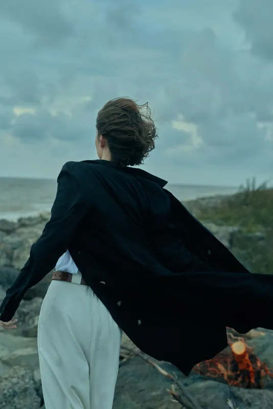 Cape worn figure on windy coastline runs along the beach with a cape billowing, flames glow nearby creating dramatic motion and bold atmosphere
