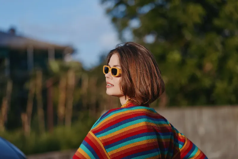 Woman, striped sweater, sunglasses, bob haircut: young woman in her late 20s wearing yellow sunglasses and a colorful striped sweater, smiling over her shoulder in warm outdoor suburban light.