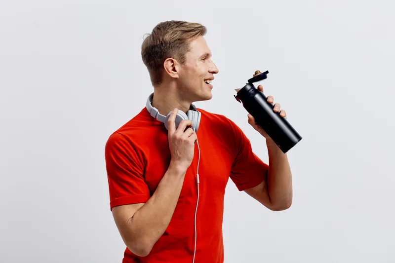 Young smiling man in red t-shirt holding a black water bottle with a lid isolated on plain white background. Casual lifestyle concept, healthy hydration, active leisure, summer day, cheerful mood, fit