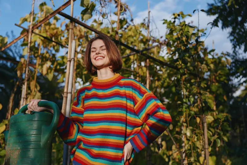 Woman in her mid 20s with short hair smiling while holding a green watering can in a sunlit outdoor garden, wearing a colorful striped sweater for casual lifestyle gardening and homegrown plants.