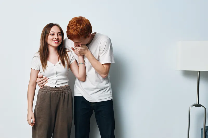 Young couple, man and woman, standing together against white wall. Man kisses woman hand, romantic gesture with casual clothes and happy facial expressions indoors.