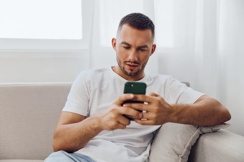 Engaged man using smartphone on a cozy couch, showcasing modern technology and comfort in a bright living space. Perfect for lifestyle and tech themes.