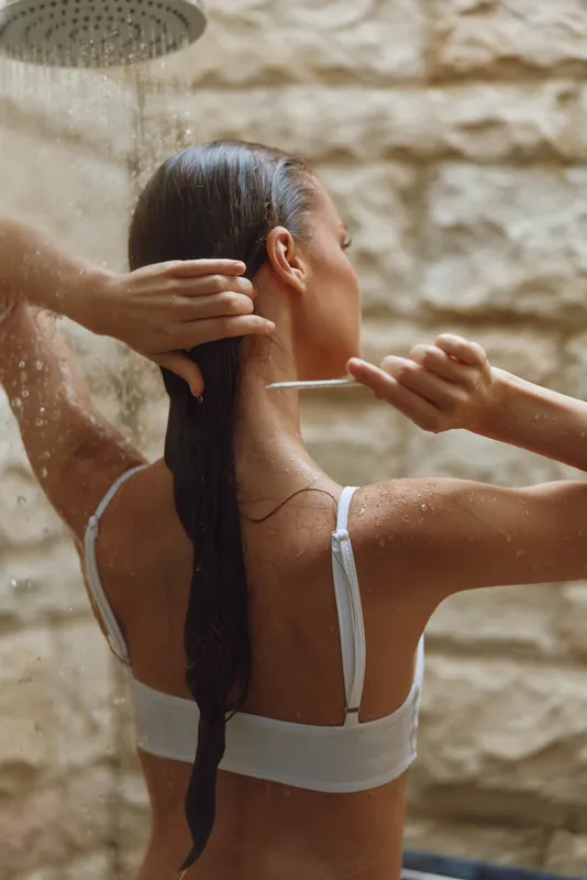 Woman washing long wet hair under outdoor rain shower with stone background, holding white comb, natural light, calm and refreshing atmosphere.