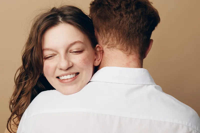 Happy young woman with closed eyes embraces man wearing white shirt. Warm intimate moment of affection and love between couple on beige background. Concept of relationship and happiness.