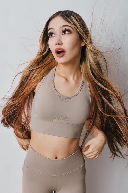 Young woman in sportswear with long hair posing and looking upward indoors. Fitness, healthy lifestyle and confident female with natural makeup standing on plain background.