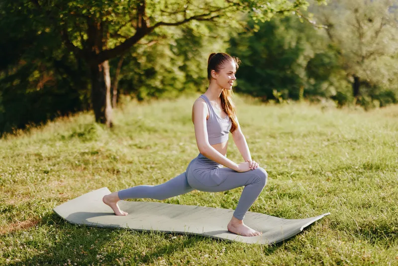Young woman practicing yoga outdoors in a serene green landscape, showcasing a calm and focused expression while wearing stylish activewear