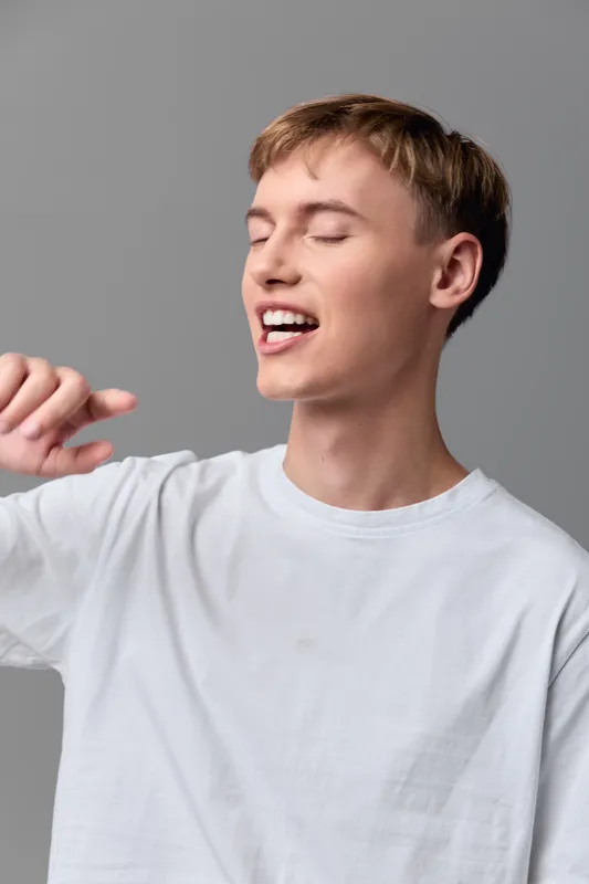 Yawning, stretching motion from a casual male in a white shirt, relaxed expression and open body language, suggesting morning routine, fatigue or refreshment in a simple studio setting
