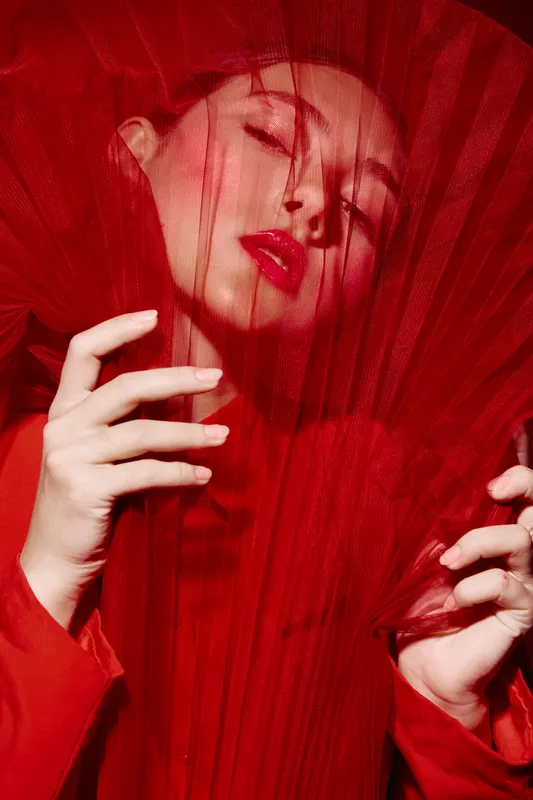 Fashion creative portrait of young woman behind red tulle fabric with vivid red lips and painted nails. Artistic beauty shot symbolizing expressive style and modern glamour.