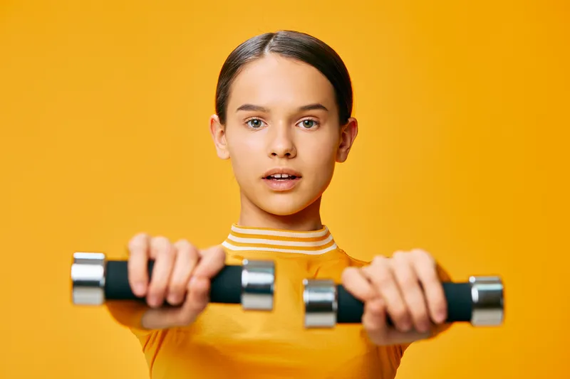 Young woman in a bright yellow top lifts dumbbells with focus, showcasing strength and fitness in a bold studio with a vivid orange backdrop.
