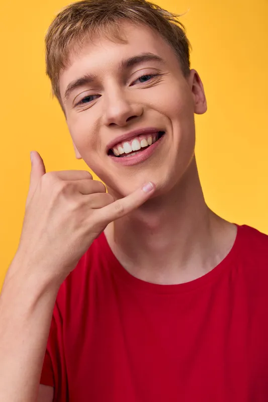 Smiling man in a red shirt makes a playful hand gesture on a bright yellow backdrop, conveying friendly energy, casual style, and a youthful approachable vibe.