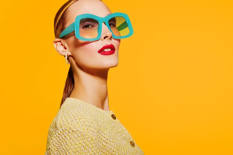 Stylish woman with large blue glasses and red lipstick poses confidently against bright yellow background. Fashion concept with bold colors and modern accessories.