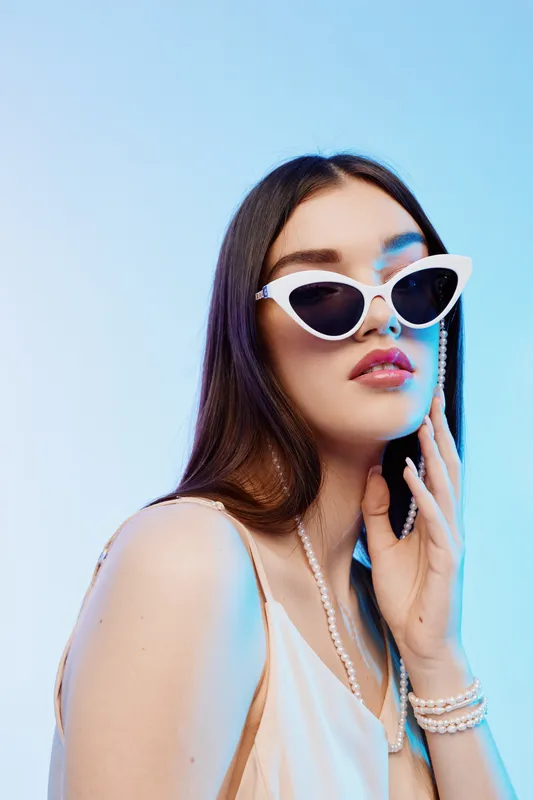 Young woman with long dark hair wearing white sunglasses and a cream dress poses with pearl accessories on a light blue background. Concept of beauty fashion and style.