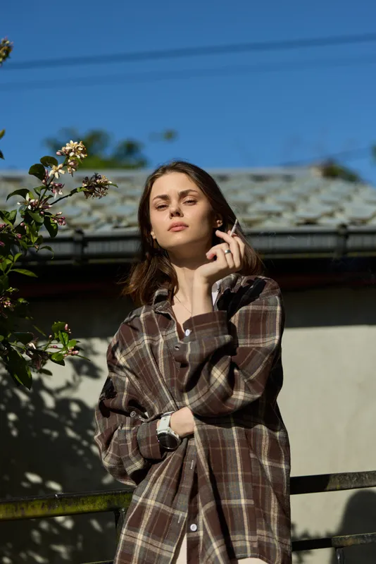 Woman lifestyle outdoor portrait in casual plaid shirt, relaxed pose under bright sky, natural sunlight, simple everyday moment, confident calm expression, candid contemporary vibe