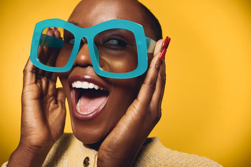 Cheerful black woman with dark skin wearing oversized blue glasses poses with open mouth on yellow background. Concept of fashion, joy, and vibrant lifestyle with bright studio lighting.