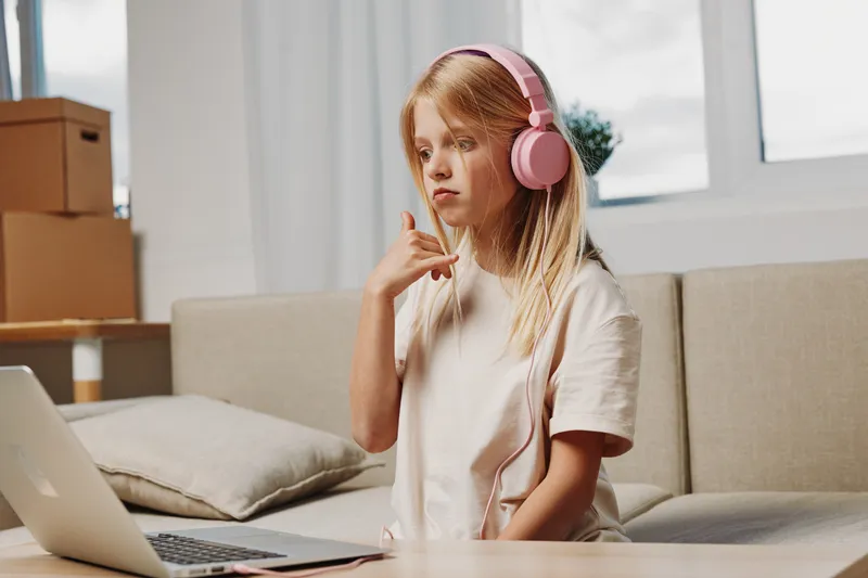 Thoughtful girl with pink headphones sits on sofa using laptop, gesturing with hand in bright living room. Concept of remote learning and communication.