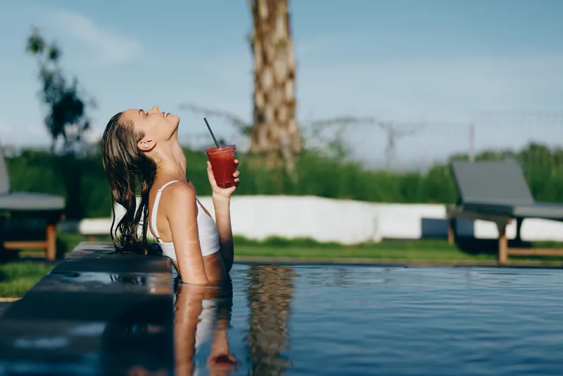 Cheerful woman outdoors by a pool, sipping a drink, head tilted back, sunlit moment, relaxed posture, vibrant poolside scene with palm trees and lounge chairs