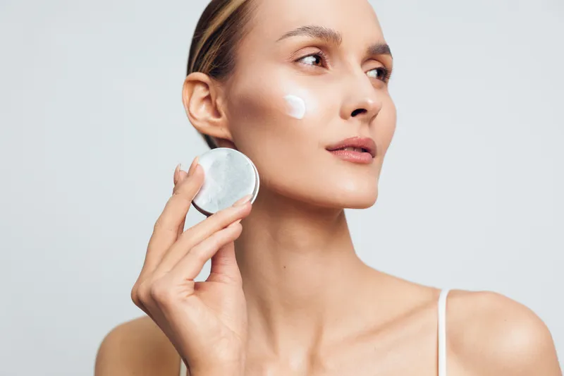 Woman skincare face cream cotton pad beauty hygiene portrait with clean studio light, she applies moisturizer to her cheek, calm expression, closeup beauty routine for skin care